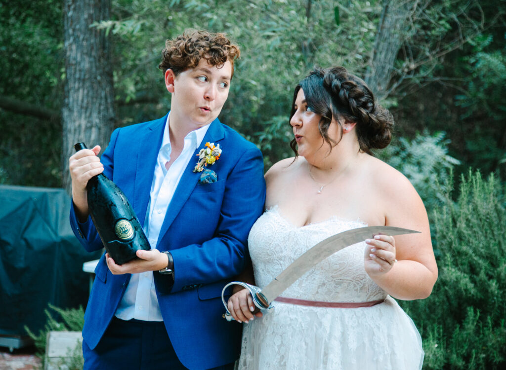 plan a stress free los angeles wedding, Brides after sabering a bottle of champagne, saber still in hand. one bride in a blue suit and the other bride in a strapless white wedding dress