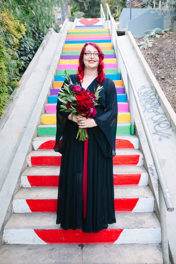 Bride with red hair in Black wedding dress with red bouquet standing on rainbow steps