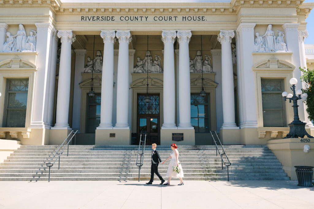 two brides walking in front of the riverside county courthouse, one bride in a black suit and one in a white dress. lesbian wedding, lgbtq+ wedding photographer