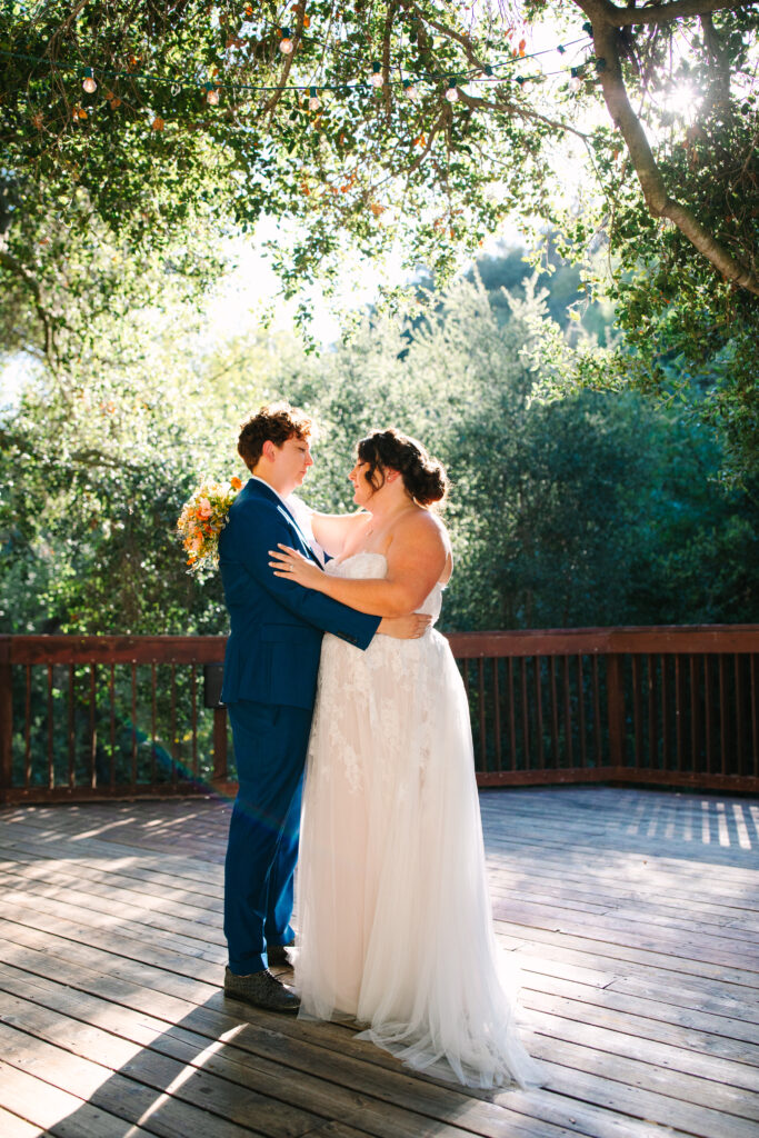 lgbtq wedding photographer, two brides backlit one bride in white strapless dress and the other bride in a blue suit. Standing below an oak tree standing and embracing looking into each other's eyes.