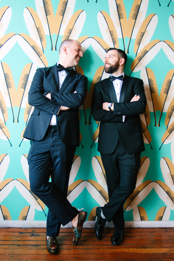 two grooms in black suits looking at each other leaning against cool palm leaf wallpaper