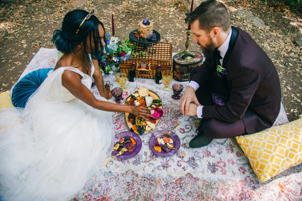 spend vs. splurge wedding, picnic in the woods bride and groom sitting on a rug and eating charcuterie