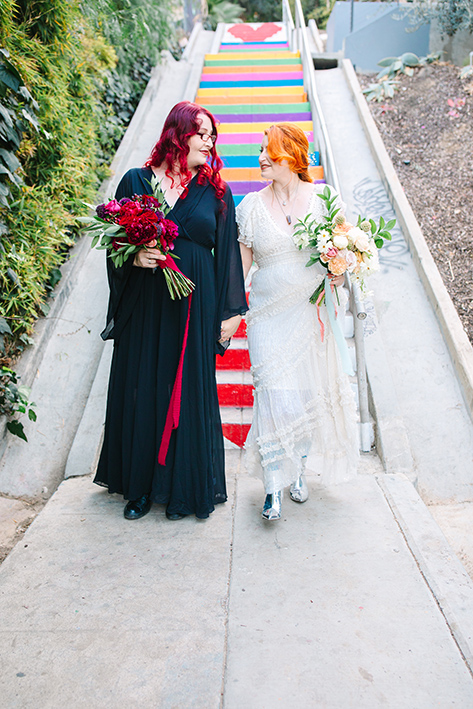 candid wedding photography, two brides one in a black dress one in a white dress walking hand in hand in front of rainbow steps in los angeles