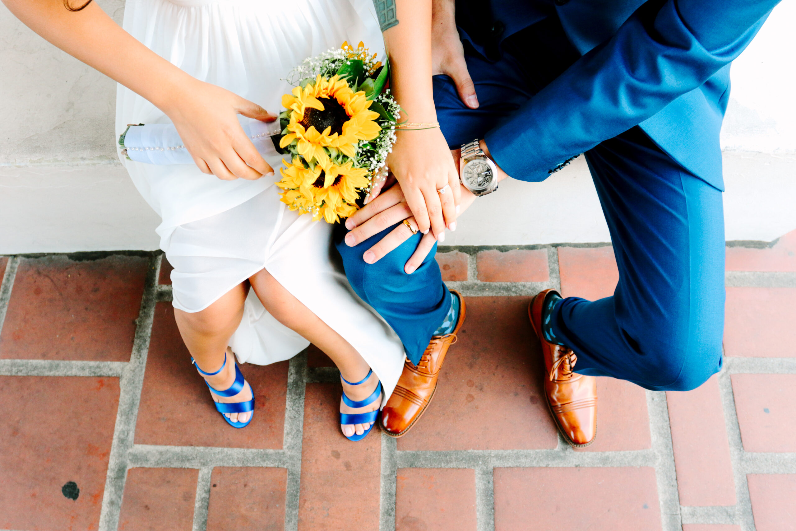 planning your wedding, bride in white and groom in blue, they are sitting and it's a shot of their laps showing off their rings, shoes and her sunflower bouquet