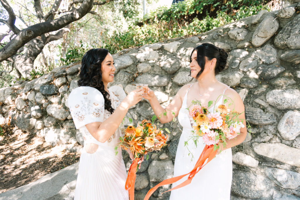 candid wedding photography, two brides in white holding their bouquets in shades of orange cheersing shots of tequila