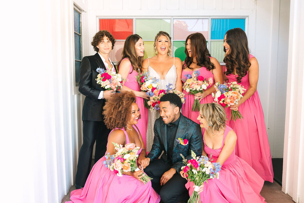 Madonna Inn Wedding Photographer, bride in white with her side of the wedding party, girls in pink and guys in black with colorful glass in the background.