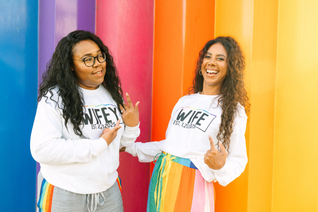 feel comfortable in front of the camera, lesbian engagement session, two women in white sweatshirts that say wife and wifey in rainbow pants in front of a rainbow wall smiling and pointing at their engagement rings.