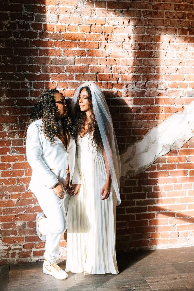 two black brides standing against a brick wall one in a white jumpsuit with a white veil and the other in a white pants suit. They are looking at each other. Lesbian wedding photographer, lgbtq wedding photographer, queer wedding photographer