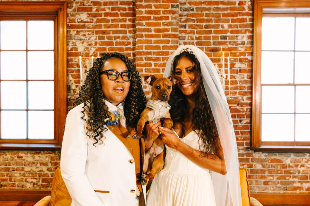 Altar Society Wedding in Long Beach, Lesbian Wedding brides in white jumpsuit and white suit holding their dog, the ringbearer for a photo